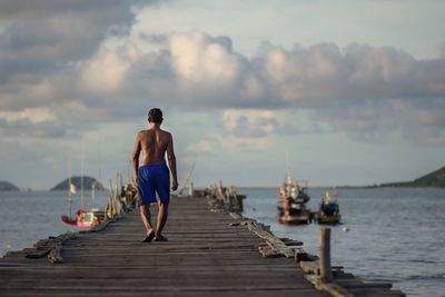 Rear view of shirtless man walking on pier over sea against sky at sunset