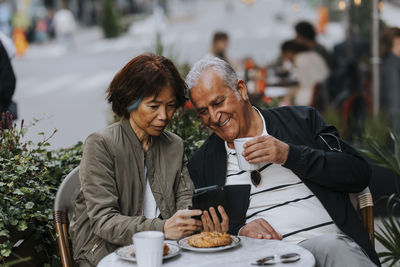 Senior woman sharing smart phone with elderly man holding coffee cup at sidewalk cafe