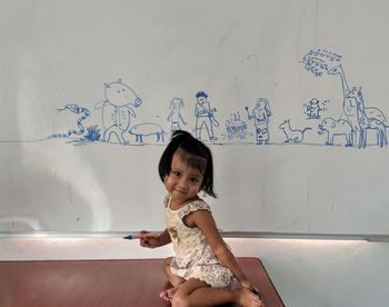 Full length portrait of girl sitting against drawings on white board
