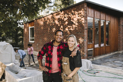Portrait of confident couple standing while children playing in background against house
