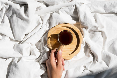 Females hand reaches for morning coffee cup in bed. woman wakes up on white bedding in sunlight.