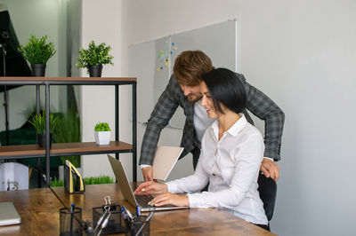 A man stands next to a woman sitting at a desk with a laptop in the office