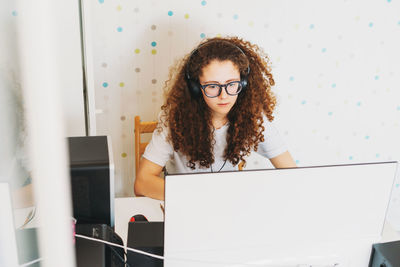 Curly hair girl teenager in glasses and headphones in white t-shirt sitting working on computer 