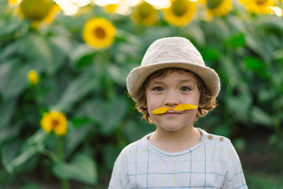 Happy little boy walking in field of sunflowers and making a mustache from sunflower petals.