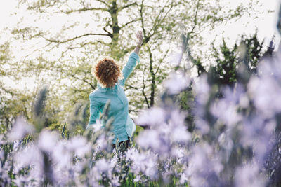 Happy woman standing with arms raised amidst flowering plants at forest