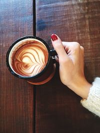 Cropped hand of woman holding cappuccino in cup on wooden table