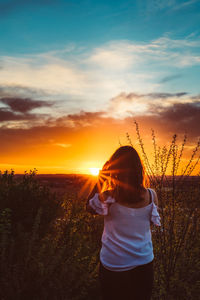 Rear view of woman standing by plants against sky during sunset