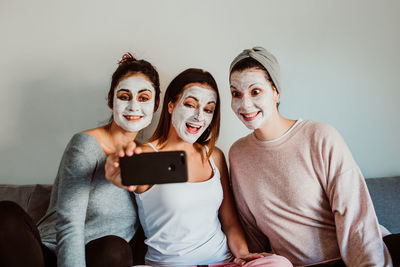 Female friends with facial masks taking selfie with against wall at home