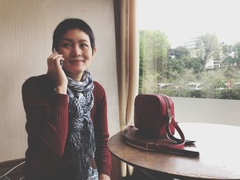 Portrait of young woman using phone while standing on window