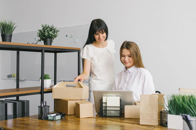 Two girls in the office are working with a laptop next to eco packages shelves with folders standing