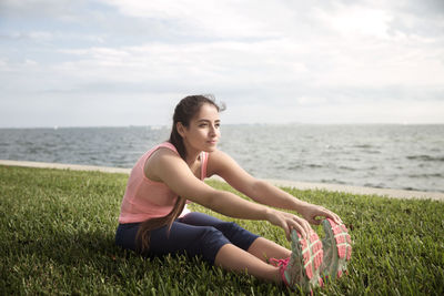 Young woman touching toes while sitting on grassy field against sea