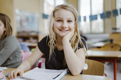 Smiling blond schoolgirl leaning on elbow while sitting at desk in classroom
