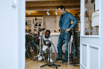 Smiling owner repairing bicycle in workshop by colleague seen through glass window