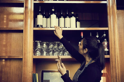 Side view of female owner with phone checking list of wine bottles while standing in cafe