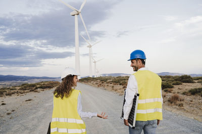 Smiling colleague gesturing at engineer holding solar panel at wind farm