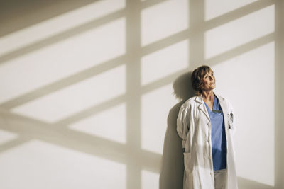 Female doctor wearing lab coat day dreaming while standing against wall with sunlight