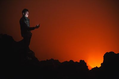 Side view of man using mobile phone while standing on silhouette rocks during sunset