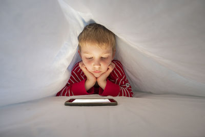 Boy using mobile phone while lying on bed under blanket