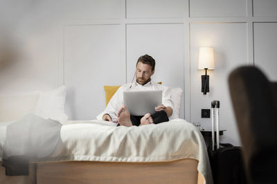 Businessman sitting on bed using laptop against wall in hotel room