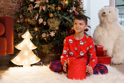 Happy boy in red pajamas sitting on the floor by the christmas tree with a gift