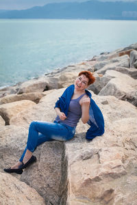 Full length portrait of smiling young woman sitting on rock at beach