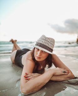 Portrait of woman wearing hat lying at beach against sky