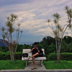 Woman sitting on chair by palm trees against sky