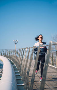 Young woman jogging on footbridge against clear sky