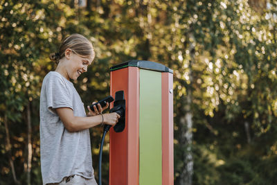 Happy boy using smart phone while holding electric plug at car charging station