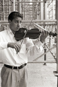 Man playing violin while standing on footpath against scaffolding