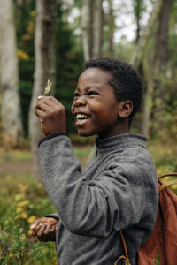 Side view of cheerful boy examining leaf during vacation in forest