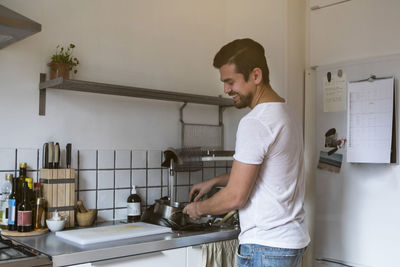 Smiling man washing utensils while standing by sink at kitchen
