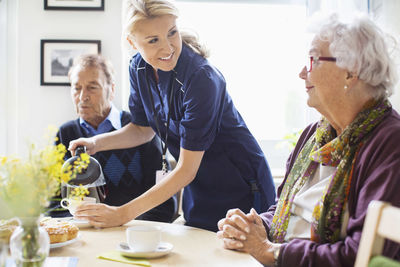Happy female caretaker serving coffee to senior people at nursing home