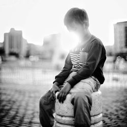 Boy sitting on concrete bollard in city during sunset