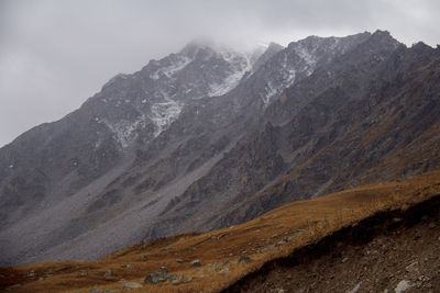 High rocky mountain with snow-capped peaks in fog against sky and clouds in bad weather in autumn