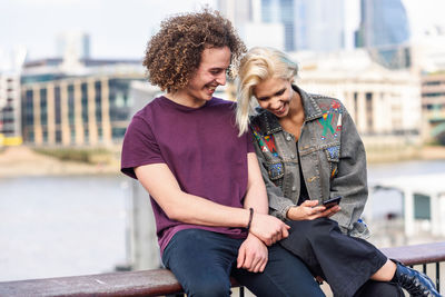 Couple using mobile phone while sitting on railing in city