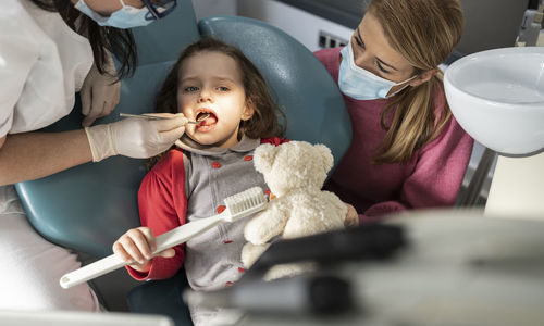 Mother in protective face mask looking at daughter examined by dentist at medical clinic