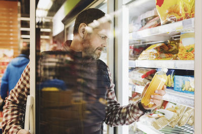 Male customer reading drink label seen through glass door at refrigerated section