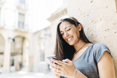 Happy young woman using mobile phone while standing against wall at city