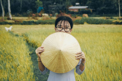 Portrait of woman holding asian style conical hat while standing on field