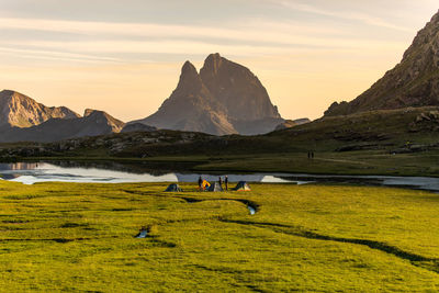 Group of traveling friends with camping tents sitting on grassy shore near calm lake reflecting mountains and enjoying spectacular sundown in summer evening