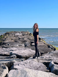 Rear view of woman standing on rocks at beach against clear sky