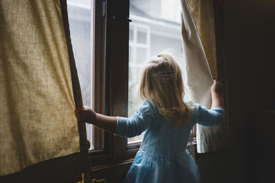 Rear view of girl looking through window while standing at home