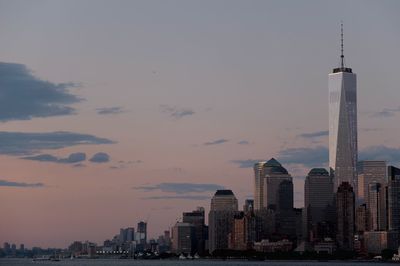 One world trade center amidst buildings in front of river against sky at dusk