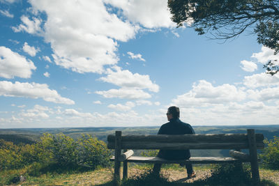 Rear view of man sitting on park bench by tree against cloudy sky during sunny day