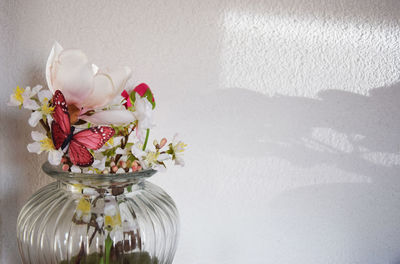 Close-up of flowers in vase with artificial butterfly against white wall