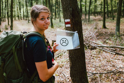 Portrait of woman showing paper with anthropomorphic face on tree trunk in forest