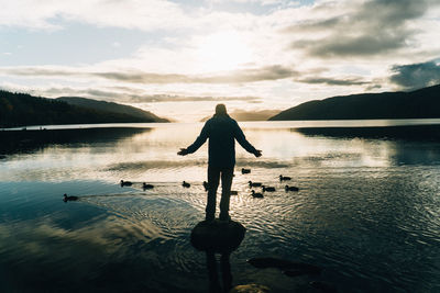 Rear view of silhouette man standing on rock by birds in lake against sky