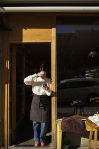 Female barista shielding eyes while using phone at doorway on sunny day