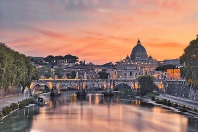 Bridge over river by buildings against sky during sunset